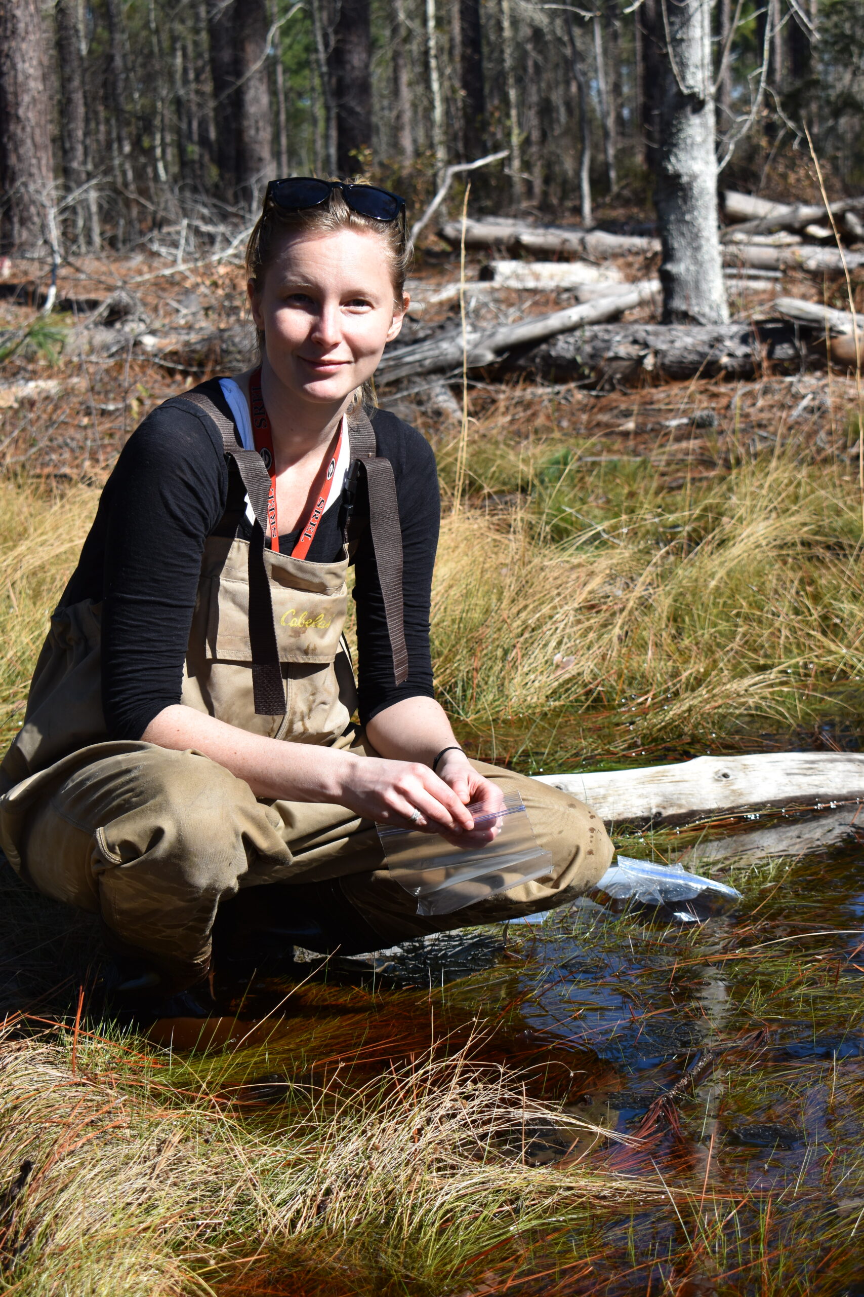 A person wearing waders and sunglasses kneels by a stream in a forested area with dense tree cover, holding a plastic bag in their hands.