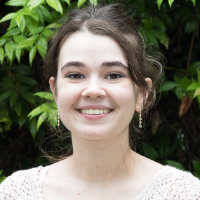 A young woman with dark hair tied back, wearing a light sweater and earrings, smiles in front of green leafy foliage—a bright moment captured among graduate students.