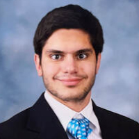 A young man with short dark hair, wearing a dark suit, white shirt, and blue patterned tie, poses in front of a blue and gray background—typical attire for graduate students preparing for professional portraits.