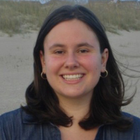 A woman with shoulder-length brown hair and hoop earrings smiles outdoors, sand and grass in the background, capturing the relaxed spirit often found among graduate students.
