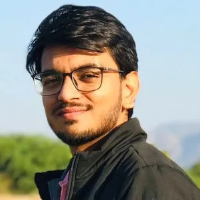 A man with dark hair and glasses, dressed in a black jacket and pink shirt, stands outdoors with greenery and a blue sky in the background, embodying the thoughtful style often seen among graduate students.