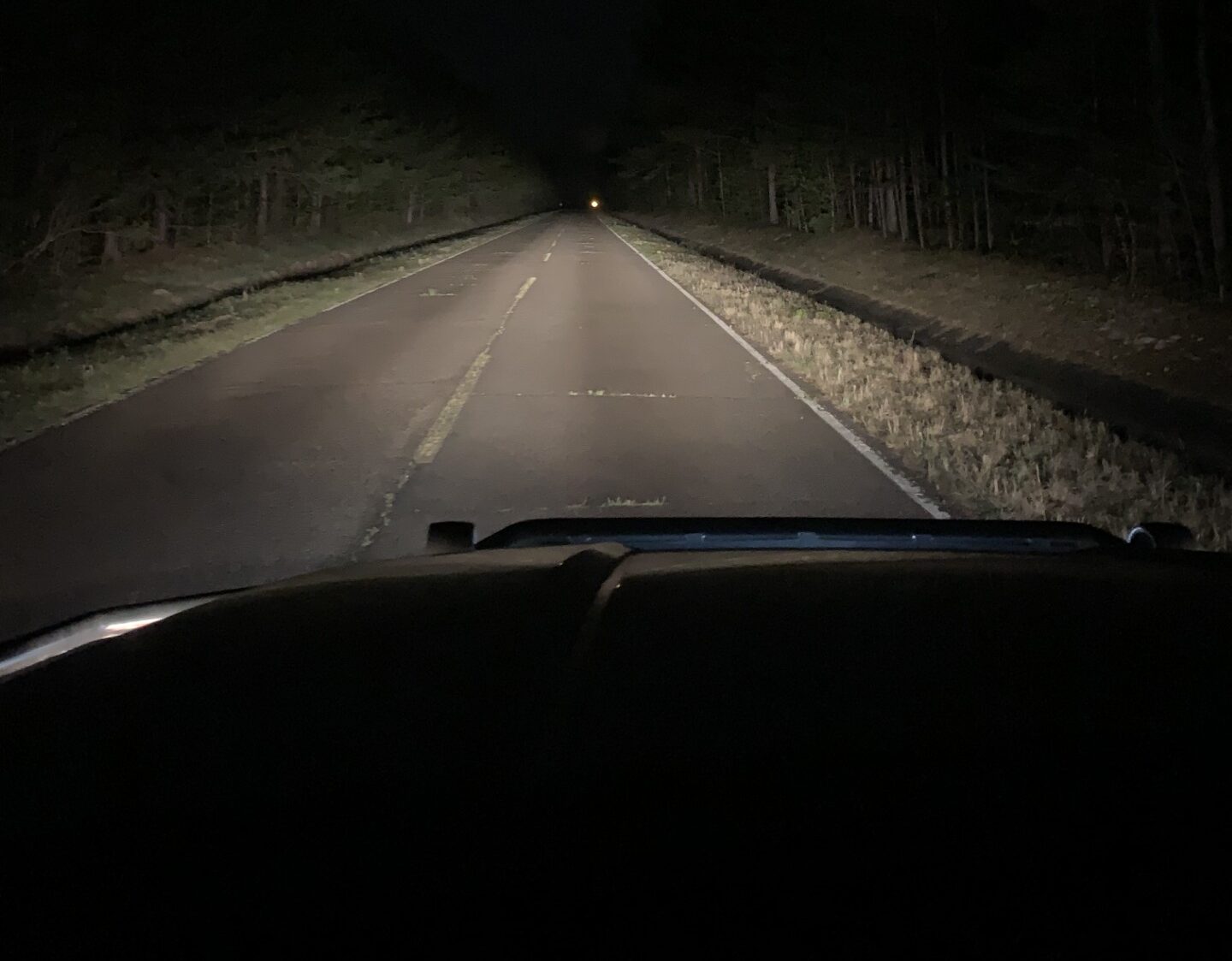 A dark, empty road stretches ahead at night, illuminated by vehicle headlights, with trees lining both sides and a single distant light visible—a reminder of the importance of road safety for every driver.