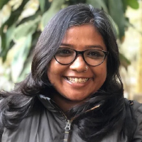 A woman with shoulder-length dark hair and glasses, likely one of the graduate students, smiles at the camera in a dark jacket. Green leaves are visible in the background.