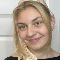 A young woman with blonde hair, possibly one of the graduate students, wears a patterned beige headscarf and black top as she smiles slightly in front of a white door.