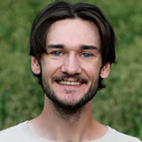 A young man with brown hair and a beard, likely one of the graduate students, is smiling at the camera while standing outdoors with greenery in the background.