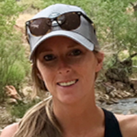 A woman, possibly one of the graduate students, wearing a gray cap and sunglasses on her head stands outdoors near trees and a creek, smiling at the camera.