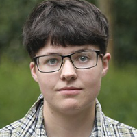 A graduate student with short dark hair and glasses, wearing a plaid shirt, stands outdoors with greenery in the background.