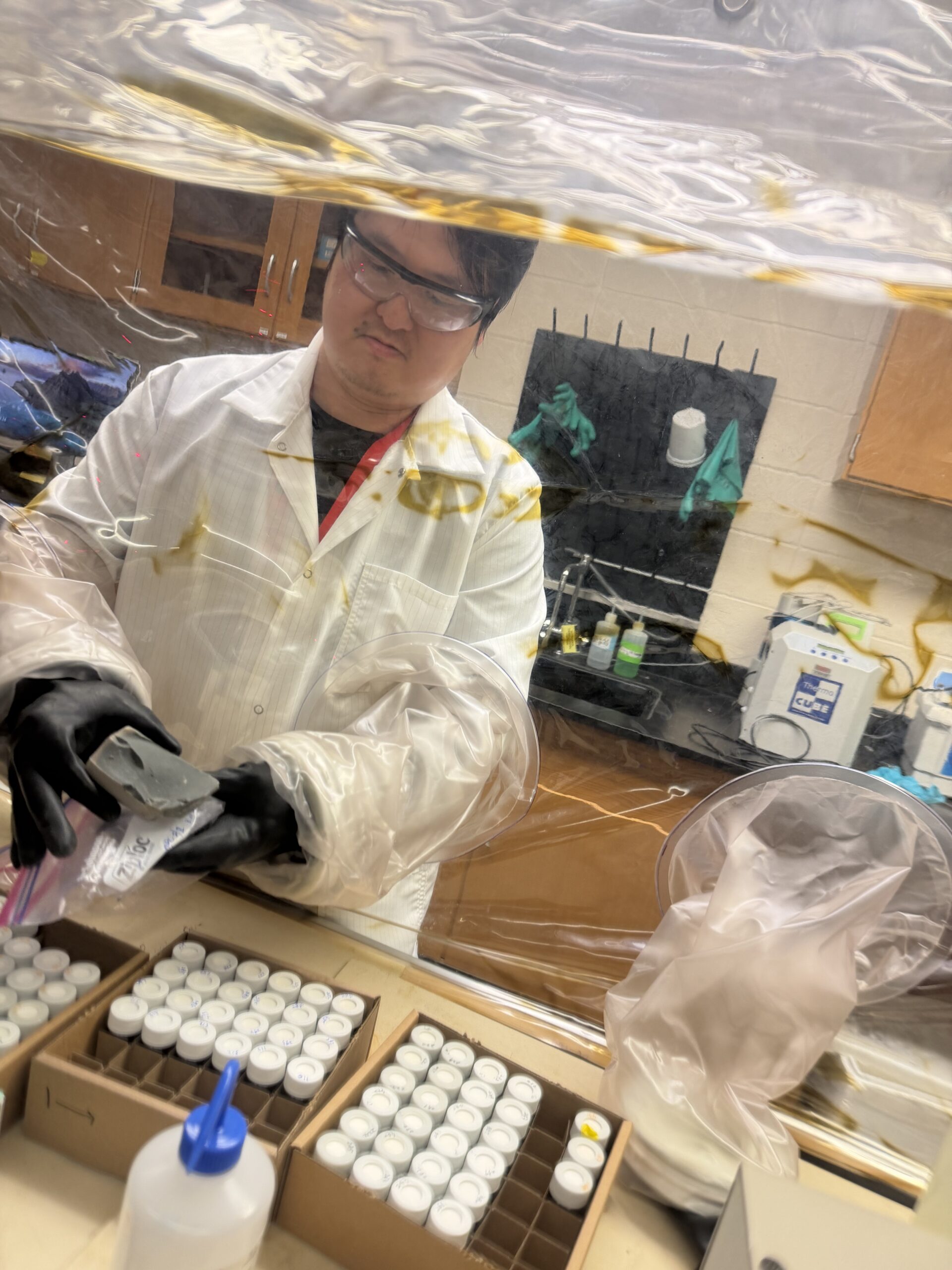 A person in a lab coat, gloves, and goggles handles samples inside a protective enclosure in a laboratory, revealing that beneath the surface of science, nothing’s set in stone. Vials and lab equipment are visible on the counter.