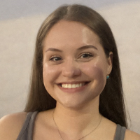 A young woman with long brown hair and a gray tank top smiles at the camera against a neutral background, embodying the confidence and optimism often seen in graduate students.