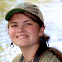 A graduate student smiles at the camera, wearing a green cap and earrings, with water blurred in the background.