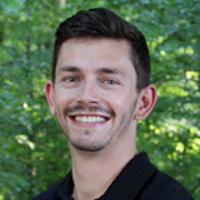 A man with short brown hair and a trimmed beard, wearing a black collared shirt, smiles in front of green trees—an approachable image often seen among graduate students.