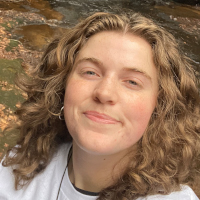 A graduate student with curly light brown hair smiles at the camera, wearing a white top, outdoors near a stream with rocks.