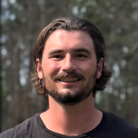 A man with medium-length dark hair and a beard is smiling outdoors, standing in front of blurred trees while wearing a black shirt—capturing the relaxed spirit often seen among graduate students.
